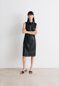 Abito senza maniche in pelle nera con bottoni frontali, due tasche sul petto e una silhouette dritta. Modello abbinato a sandali con tacco neri.