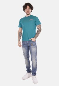 T-shirt teal a maniche corte con collo rotondo. Abbinato a jeans azzurri strappati e sneakers bianche. Dettaglio logo semplice sulla maglietta.