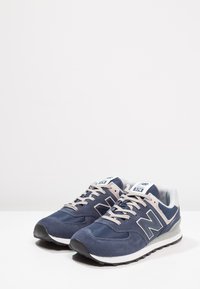 Sötétkék sportcipők velúr és hálós anyagból, fehér talppal, szürke részletekkel és bézs fűzőkkel, a New Balance logójával.