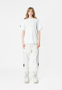 Joven de pie de frente, vistiendo una camiseta blanca holgada, pantalones blancos con diseños de alas y botas blancas con cordones sobre un fondo liso.