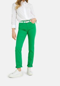 Pantalons en coton vert à coupe slim, avec des ourlets roulés et une ceinture en cuir blanc. Portés avec une chemise blanche à col et des baskets blanches.