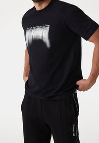 Černé bavlněné tričko s velkým, opotřebeným bílým logem "GUESS" na hrudi. Krátké rukávy a kulatý výstřih.