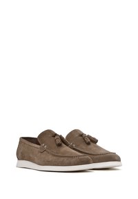 Zapatos de ante marrón de tipo slip-on con una suela de goma blanca, que presentan borlas decorativas y detalles de costura en la parte superior.