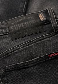 Vaqueros de mezclilla negra con una tela texturizada, que presentan una etiqueta de cuero prominente que dice "SUPERDRY", y una pequeña etiqueta roja con el nombre de la marca.