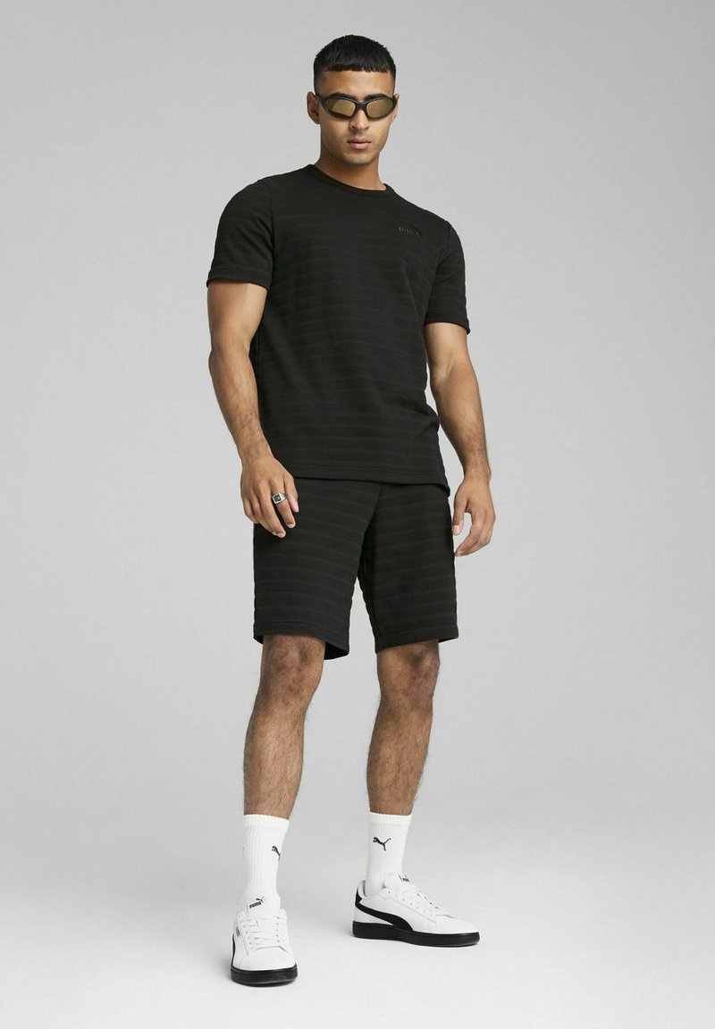 Sort stribet kortærmet skjorte parret med matchende shorts. Hvide sneakers med sorte detaljer og hvide sokker. Simpel, atletisk design.