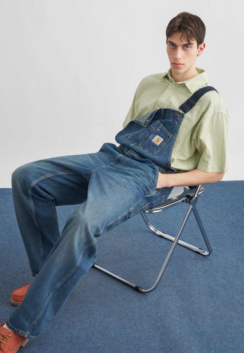 Junger Mann in grünem Hemd und blauer Jeans-Latzhose, der sich auf einem Metallklappstuhl auf einem blau teppichbedeckten Boden zurücklehnt.