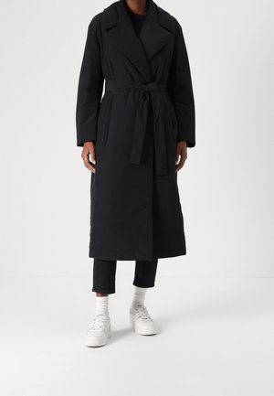Personne portant un long manteau noir ceinturé, un pantalon noir court, des chaussettes blanches et des baskets blanches sur un fond clair uni.