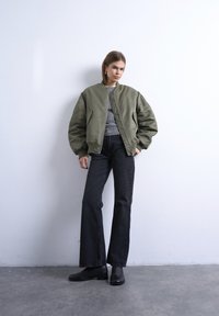 Femme debout contre un mur blanc, portant une veste bomber oversize vert olive, un t-shirt gris, un jean noir à jambes larges et des chaussures noires.