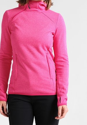 Frau trägt eine leuchtend pinke Fleecejacke mit Reißverschluss und Daumenlöchern, kombiniert mit schwarzen Hosen, vor einem schlichten hellen Hintergrund.