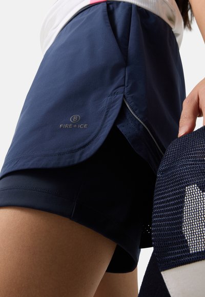 Nahaufnahme einer Person, die marineblaue Sportshorts mit dem Logo „FIRE + ICE“ trägt und eine marineblaue Netzkappe in der rechten Hand hält.