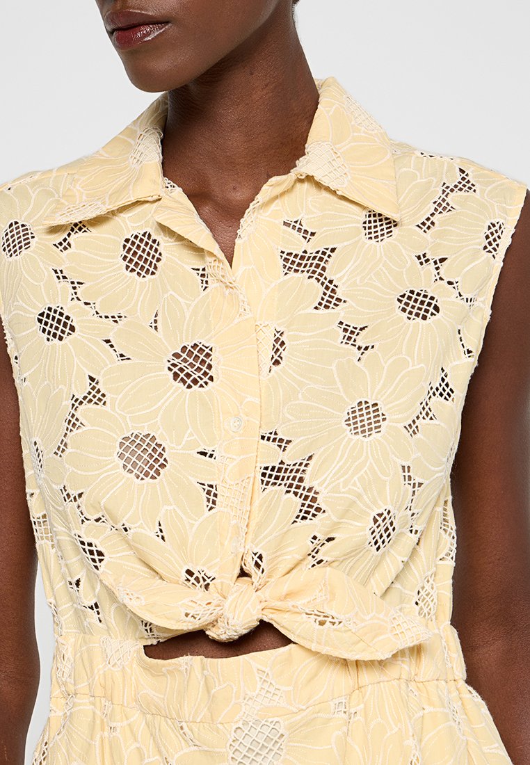 Camiseta sin mangas de color amarillo claro con diseño de encaje floral, cierre de botones y detalle de nudo en el frente con un patrón texturizado.