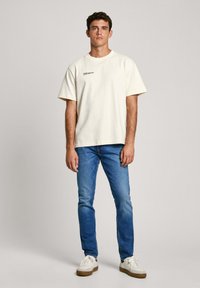 Camiseta de algodón crema de mangas cortas, con escote redondeado y logotipo negro impreso en el pecho. Lleva puesta con jeans de mezclilla azules y zapatillas blancas.