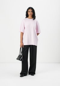 Světle růžové oversized tričko s krátkým rukávem, spárované s černými širokými kalhotami. Modelka drží malou černou kabelku a má na sobě černé boty.