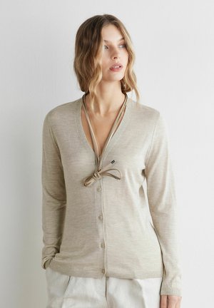 Frau mit welligem blondem Haar, die eine beigerfarbene, geknöpfte Strickjacke und lockere weiße Hosen trägt und seitlich gegen einen schlichten weißen Hintergrund schaut.