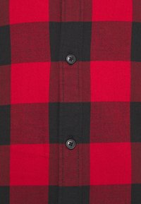 Camicia a scacchi rossa e nera in flanella, con chiusura a bottoni e texture morbida. Quattro bottoni prominenti visibili.