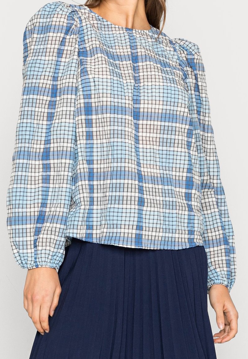 Niebieska i biała bluzka w kratkę z długimi bufiastymi rękawami, marszczonym dekoltem i elastycznymi mankietami, w połączeniu z plisowaną granatową spódnicą.
