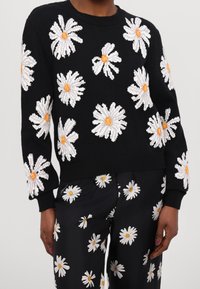 Svart stickad tröja med vit och gul tusensköna broderi. Rund halsringning och ribbade muddar. Matchas med svarta byxor med liknande blommönster.