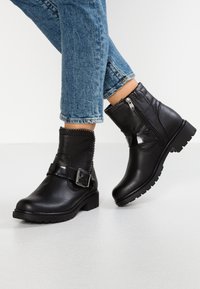Bottines en cuir noir avec une sangle et des détails de boucle, dotées d'une fermeture éclair sur le côté, d'une tige texturée avec des coutures et d'une semelle en caoutchouc épaisse.