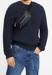 Pull en maille bleu marine à texture épaisse, assorti à un jean bleu. Sac banane noir à motifs porté en bandoulière avec deux fermetures éclair.