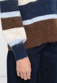 Pull en tricot avec des rayures horizontales marron, crème, marine et bleu clair. Texture douce avec des poignets côtelés et une coupe décontractée.