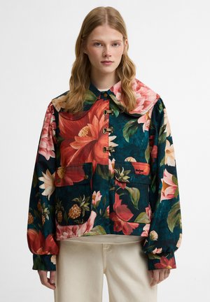 Νεαρή γυναίκα φοράει σκούρο φλοράλ μπουφάν με μεγάλα κόκκινα, ροζ και λευκά λουλούδια, μπροστινές τσέπες και κούμπωμα με κουμπιά, σε συνδυασμό με ανοιχτόχρωμο παντελόνι.