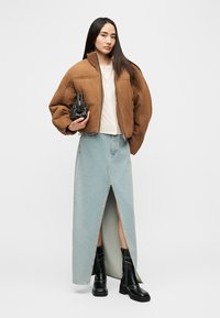 Veste bomber matelassée marron, haut crème, jupe maxi en denim bleu clair avec fente avant, et bottes noires à plateforme montant jusqu'aux genoux. Petit sac à main noir.