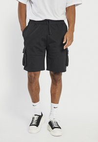 Homme portant un short cargo noir avec des poches avant, des chaussettes blanches Nike, et des baskets noir et blanc, debout devant un fond blanc.