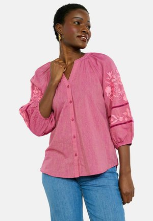 Frau in rosa Bluse mit Knöpfen und floraler Stickerei auf den Puffärmeln, trägt blaue Jeans, schaut seitlich, Hand nahe am Kragen.