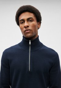 Νεαρός άνδρας με κοντά σγουρά μαλλιά, φοράει ένα ναυτικό μπλε φούτερ με φερμουάρ που είναι τραβηγμένο μέχρι τη μέση, με ουδέτερο φόντο.