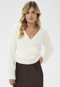 Weißer, strukturierter Wickelpullover mit V-Ausschnitt und langen Ärmeln, kombiniert mit einem dunkelbraunen Rock. Weiches und plüschiges Material.