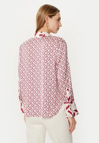 Blusa a maniche lunghe in tessuto stampato rosso e bianco. Presenta un colletto alto e bottoni decorativi sulle maniche. Texture liscia, orlo arrotondato.