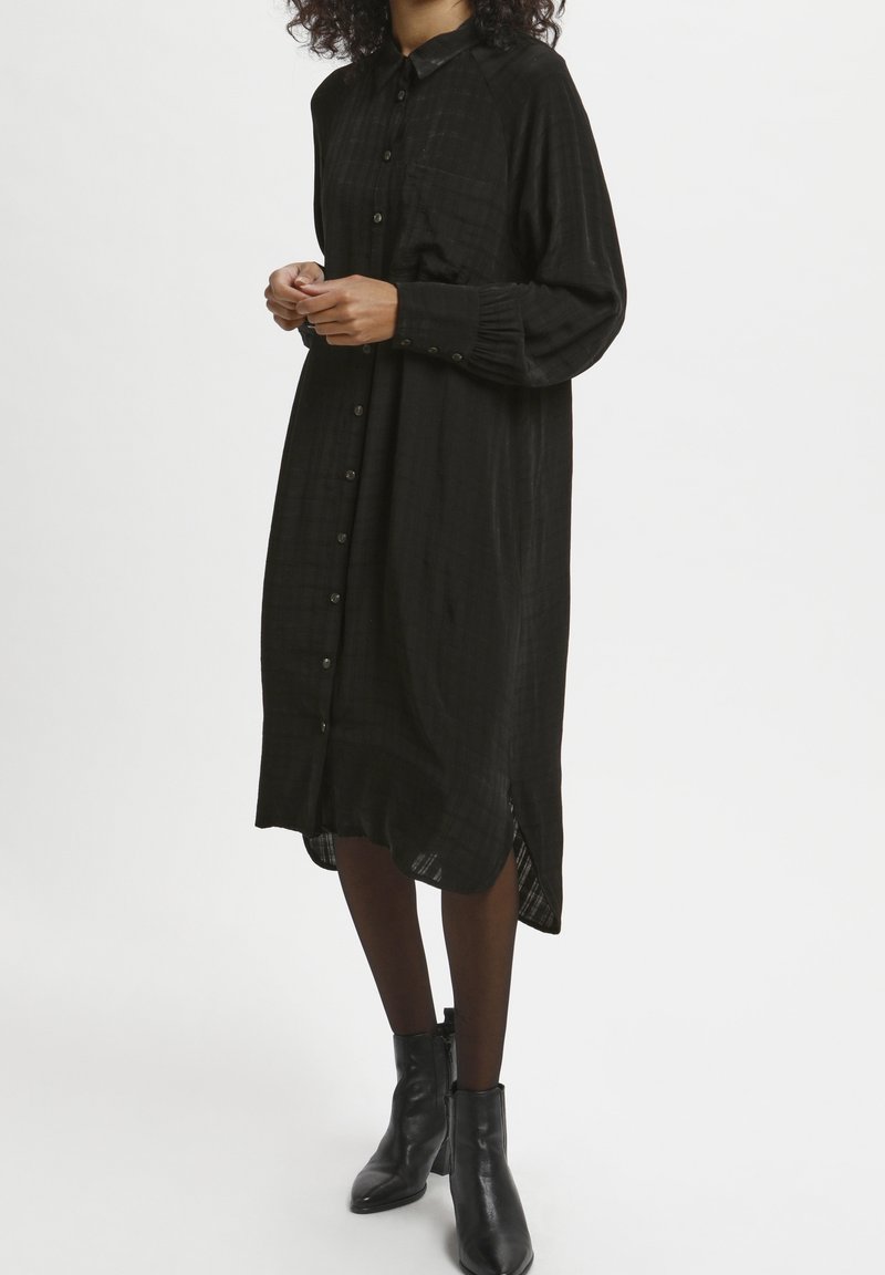 Femme debout portant une longue robe noire à boutons avec des manches longues, des collants noirs et des bottines noires sur un fond blanc.