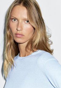 Maglione azzurro chiaro lavorato a maglia con scollo rotondo. Il modello ha capelli ondulati di lunghezza spalle e un'espressione neutra. Tessuto testurizzato.