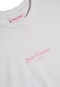 Balta medvilninė marškinėlių su apvalia apykakle, ant priekio esantis šviesiai rožinis "Juicy Couture" logotipas ir "SPORT" tekstas, lygus paviršius.