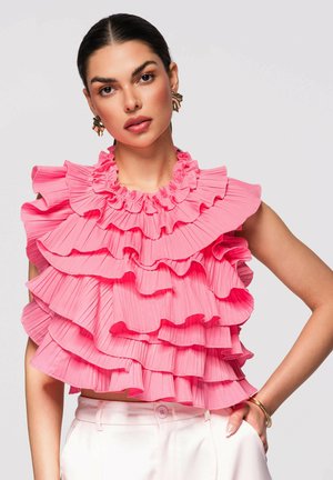 Femme aux cheveux foncés portant un haut sans manches rose à volants hauts et un pantalon blanc, posant devant un fond gris clair uni.