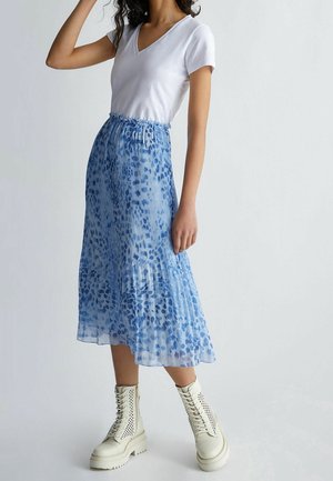 Mujer con camiseta blanca de manga corta y escote en V, falda midi plisada azul con patrón abstracto y botas blancas con cordones hasta el tobillo, de pie contra una pared blanca.
