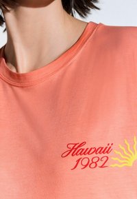Camiseta de color coral con cuello redondo, que presenta un bordado rojo de "Hawái 1982" y un gráfico de sol amarillo en la parte izquierda del pecho. Textura suave.