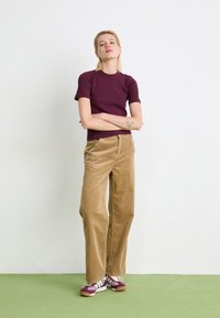 T-shirt aderente color bordeaux, pantaloni a gamba larga in corduroy color sabbia e sneakers multicolor. Tessuto morbido, design casual, con cuciture visibili e dettagli del logo.