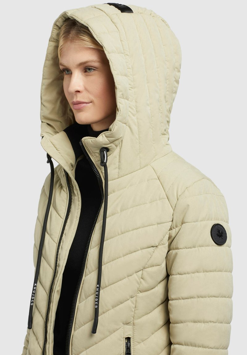 Hellbeige gesteppte Jacke mit großer Kapuze, schwarzen Kordelzügen und schwarzem Akzentknopf. Mit Zickzacknähten und einem Reißverschluss.