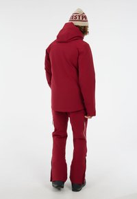 Rode winterjas en broek, met een capuchon, een gladde textuur en zijzakken met rits. Het model draagt een crèmekleurige beanie met rode letters.