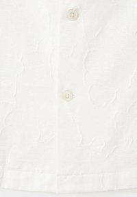 Gros plan sur un tissu blanc texturé avec deux petits boutons ronds assortis et un motif floral subtil.