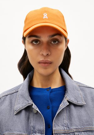 Orangene Baseballmütze mit einem weißen bestickten Logo. Die Mütze hat einen gebogenen Schirm und eine textile Struktur.getragen mit einem blauen Strickoberteil und einer grauen Denimjacke.