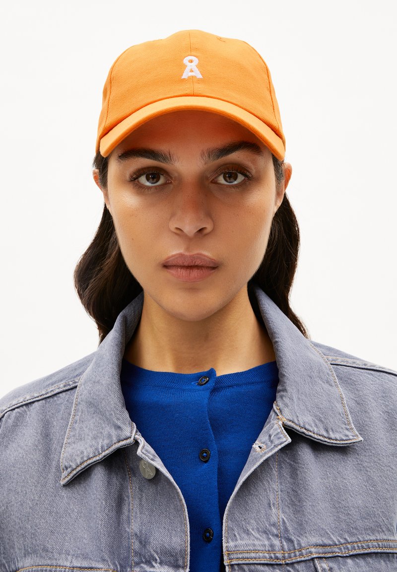 Orangene Baseballmütze mit einem weißen bestickten Logo. Die Mütze hat einen gebogenen Schirm und eine textile Struktur.getragen mit einem blauen Strickoberteil und einer grauen Denimjacke.