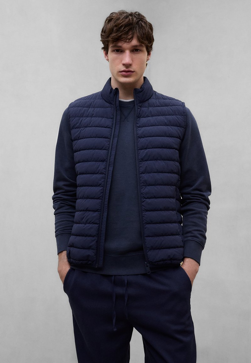 Jeune homme debout, les mains dans les poches, portant une veste matelassée bleu marine sur un sweat-shirt bleu marine et un pantalon assorti, sur un fond gris uni.