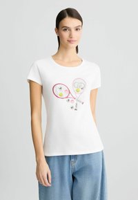 T-shirt en coton blanc présentant un graphique de deux raquettes de tennis roses et de balles de tennis colorées. Manches courtes et encolure ronde.