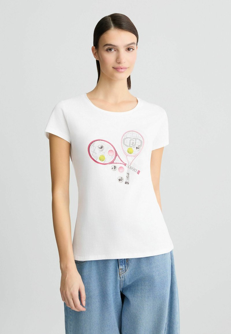T-shirt en coton blanc présentant un graphique de deux raquettes de tennis roses et de balles de tennis colorées. Manches courtes et encolure ronde.