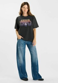 Camiseta negra de gran tamaño con texto retro colorido, combinada con jeans de pierna ancha azules y zapatos planos negros. Tejido suave y corte relajado.