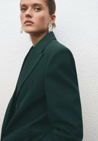 Ciemnozielona, oversize'owa marynarka z klapą typu notch i pojedynczą kieszenią, zestawiona z dużymi przezroczystymi kolczykami, na tle teksturowanej białej ściany.