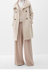 Cappotto doppiopetto a spina di pesce bianco sporco sopra un top beige con scollo a V e pantaloni a gamba larga indossato da una persona contro uno sfondo neutro.