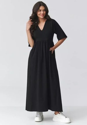 Kobieta o długich ciemnych włosach, ubrana w czarną sukienkę maxi z dekoltem w serek i rękawami do łokcia oraz białe platformowe buty sportowe, stoi z jedną ręką w kieszeni.